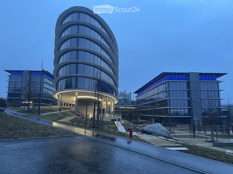Modern office complex with a prominent curved glass tower, illuminated at dusk, surrounded by contemporary buildings and landscaped grounds.