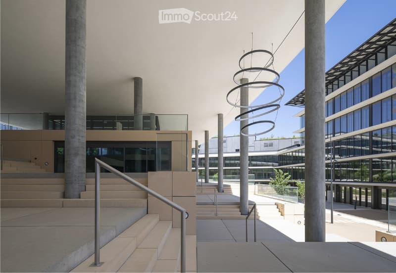 Modern building entrance with concrete steps, railings, and a glass-walled upper level. The area features large concrete columns and a contemporary sculpture in the center.