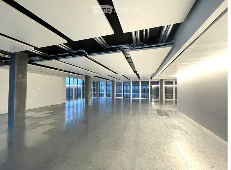 Empty room with polished concrete floor, white walls, large windows, and exposed ceiling with industrial-style lighting and ventilation ducts.