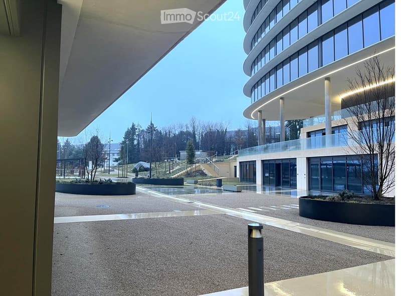 Modern building with curved glass facade, raised ground floor, and landscaped surroundings.