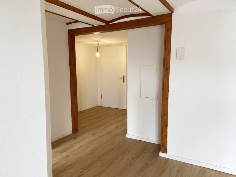 Empty room with wooden flooring, white walls, and a single door leading to another space. The room has exposed wooden beams on the ceiling and a small light fixture hanging from the ceiling.