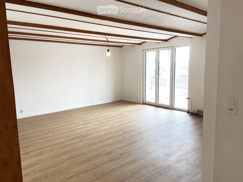 Empty room with wooden flooring, white walls, and a large sliding glass door leading to a balcony or outdoor area. Exposed wooden beams on the ceiling and a single hanging light fixture.
