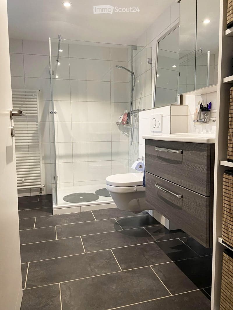 Modern bathroom with dark tiled floor, white tiled shower area, toilet, sink with mirrored cabinet, and built-in storage cabinet.