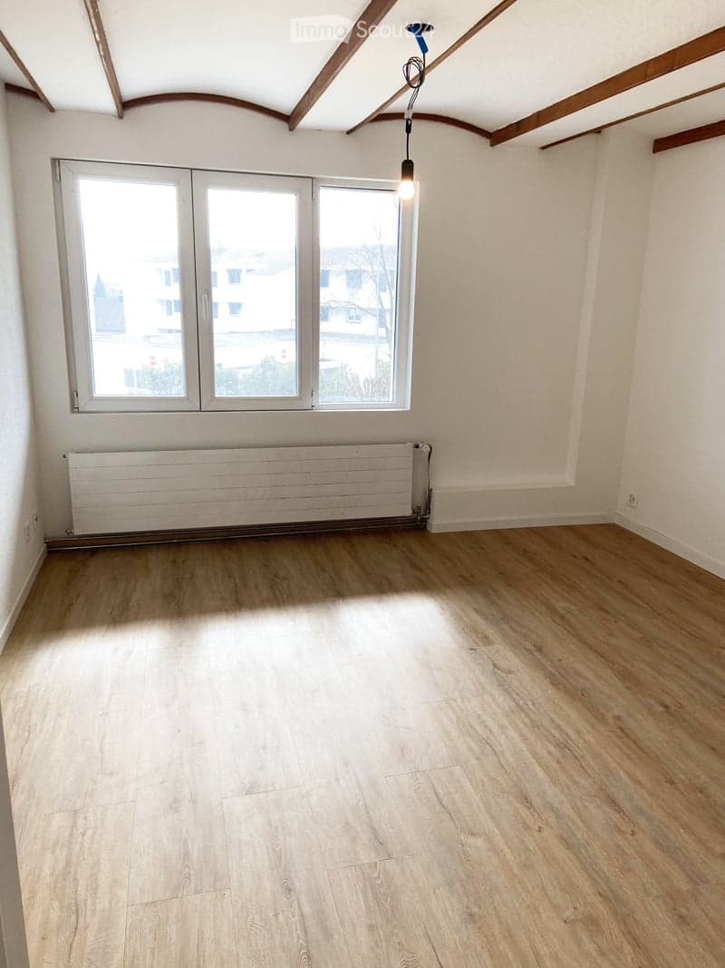 Empty room with wooden flooring, white walls, large window, radiator, and exposed wooden ceiling beams.