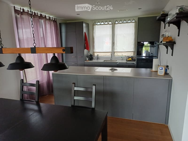 Modern kitchen with grey cabinets, a central island, a black dining table, and two chairs. Features a sink, faucet, oven, and a hanging light fixture.