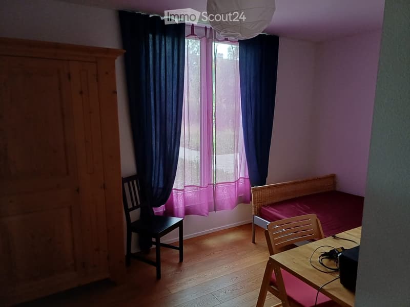 A small, cozy room with wooden flooring, a single bed with a red cover, a small table with a chair, and a wardrobe. The room has large windows with dark curtains and a hanging light fixture.