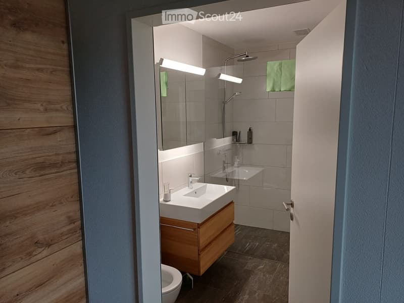 Modern bathroom with a white sink, wooden cabinet, mirror, shower, and green towels.