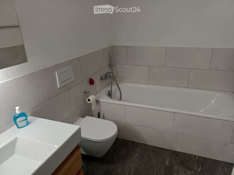 Modern bathroom with a white toilet, bathtub, sink, and mirror. Grey tiled walls and dark grey tiled floor.