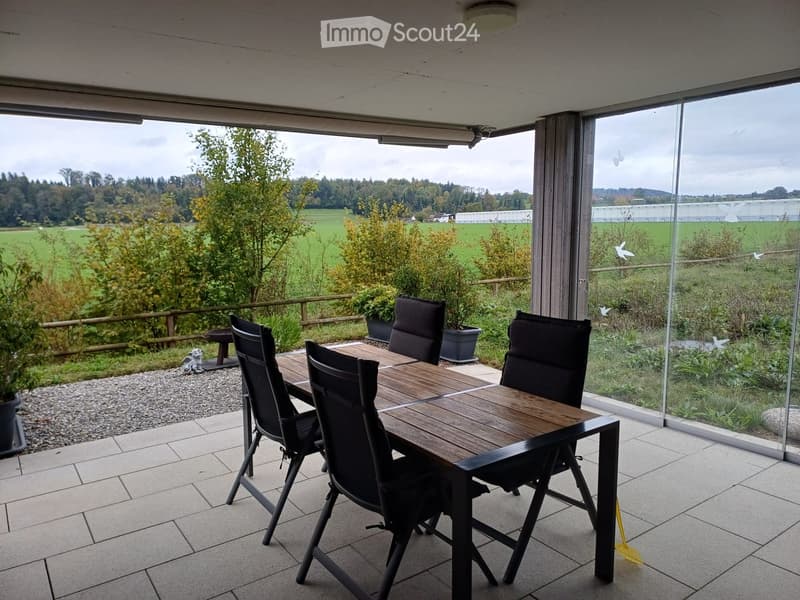 Balcony with a wooden dining table and black chairs, overlooking a green field and trees.