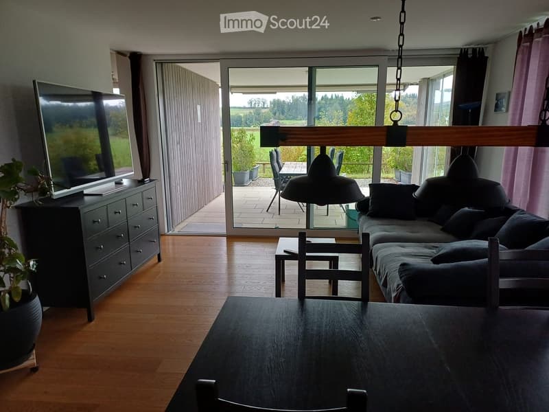 Bright living room with wooden flooring, large sliding glass doors leading to a patio, a grey sectional sofa, coffee table, dining table, and a TV on a cabinet.