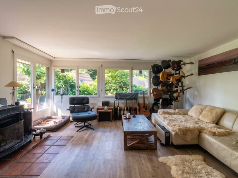 Bright living room with large windows, a fireplace, a cream-colored sectional sofa, a wooden coffee table, a black leather chair, and several guitars displayed on a rack.