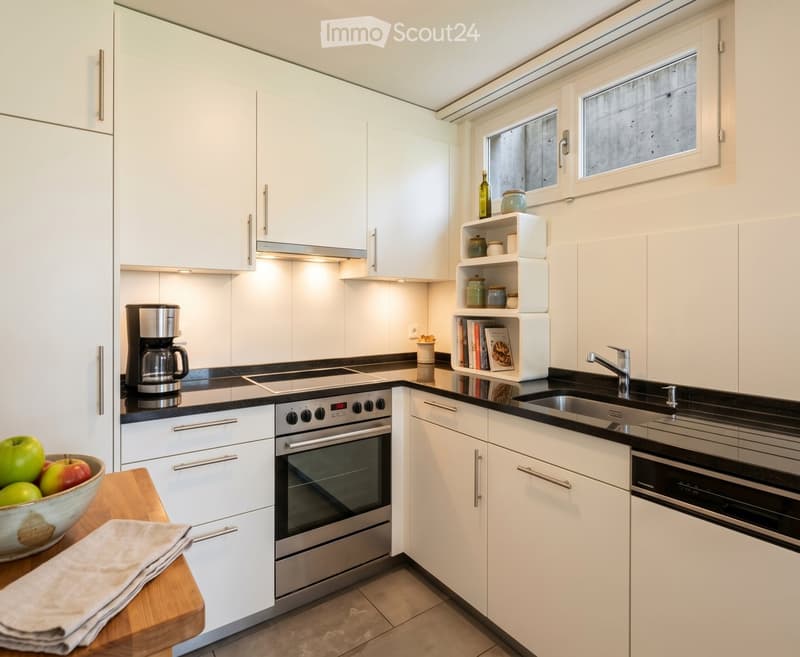 Modern kitchen with white cabinets, stainless steel appliances, and a black countertop. Features include a coffee maker, oven, sink, and a small wooden table with a bowl of fruit and a cloth.