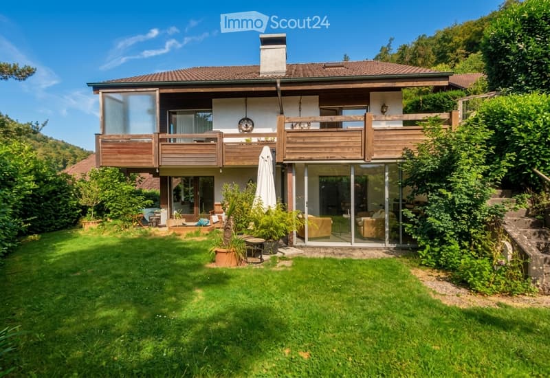 Two-story house with a tiled roof, wooden balconies, large windows, and a well-maintained garden with patio furniture.