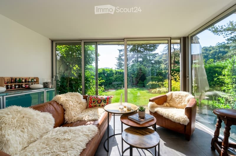 Modern living room with large glass windows offering a view of a green garden. Features a brown leather sofa with fur throws, a matching armchair, wooden tables, and decorative items.