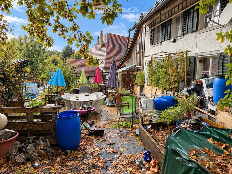 A garden area with a patio, outdoor furniture, potted plants, and various storage items, including barrels and a covered object. The space is surrounded by trees and has a wooden fence.