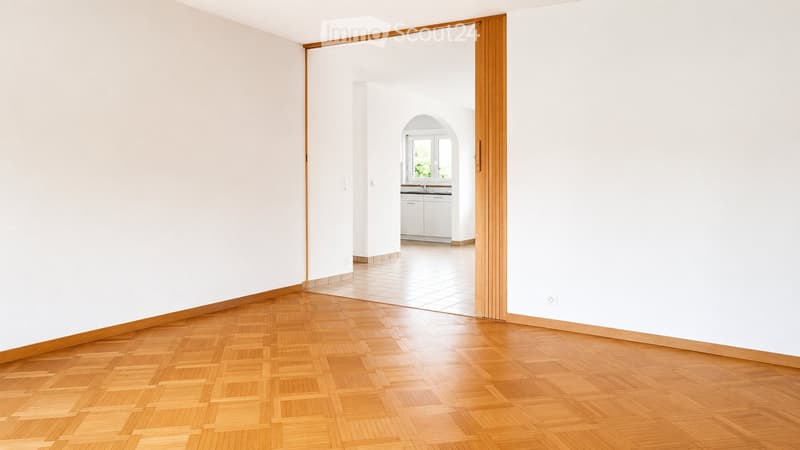 Empty hallway with wooden flooring and white walls, leading to a small kitchen area with white cabinets and a sink.