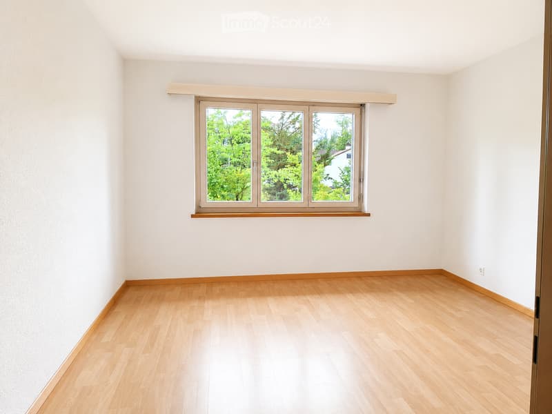 Empty room with light wooden flooring, white walls, and a large window with a view of greenery outside.
