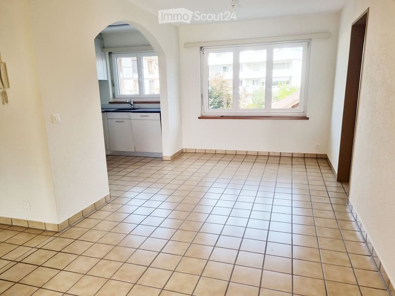 Empty room with tiled floor, white walls, and a small kitchen area visible through an arched doorway. The kitchen has white cabinets, a sink, and a window.
