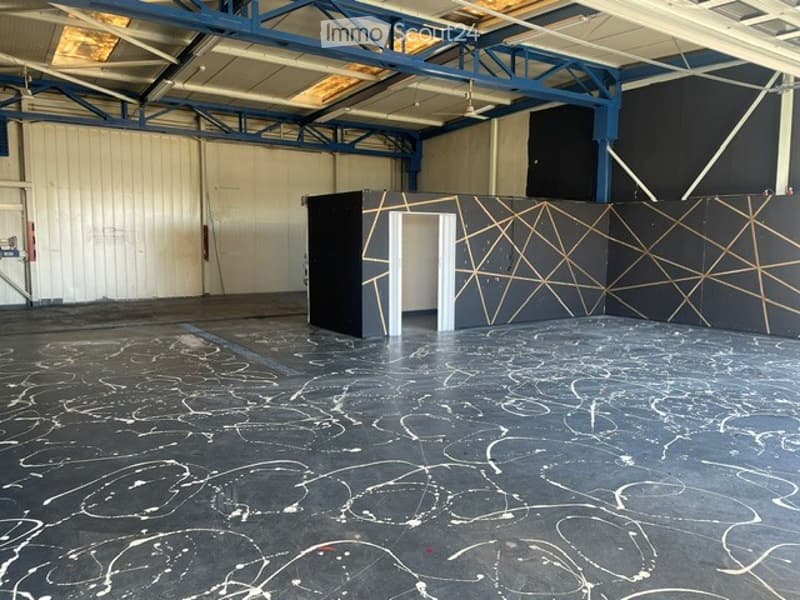 Empty industrial-style room with a high ceiling, metal beams, and a large open space. The floor is painted with abstract white markings, and there is a small black partition with a geometric design and a doorway leading to another area.