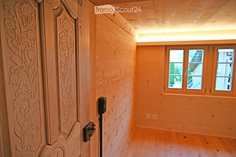 Wooden hallway with light wooden walls and floor, featuring a decorative wooden door on the left and windows at the end.