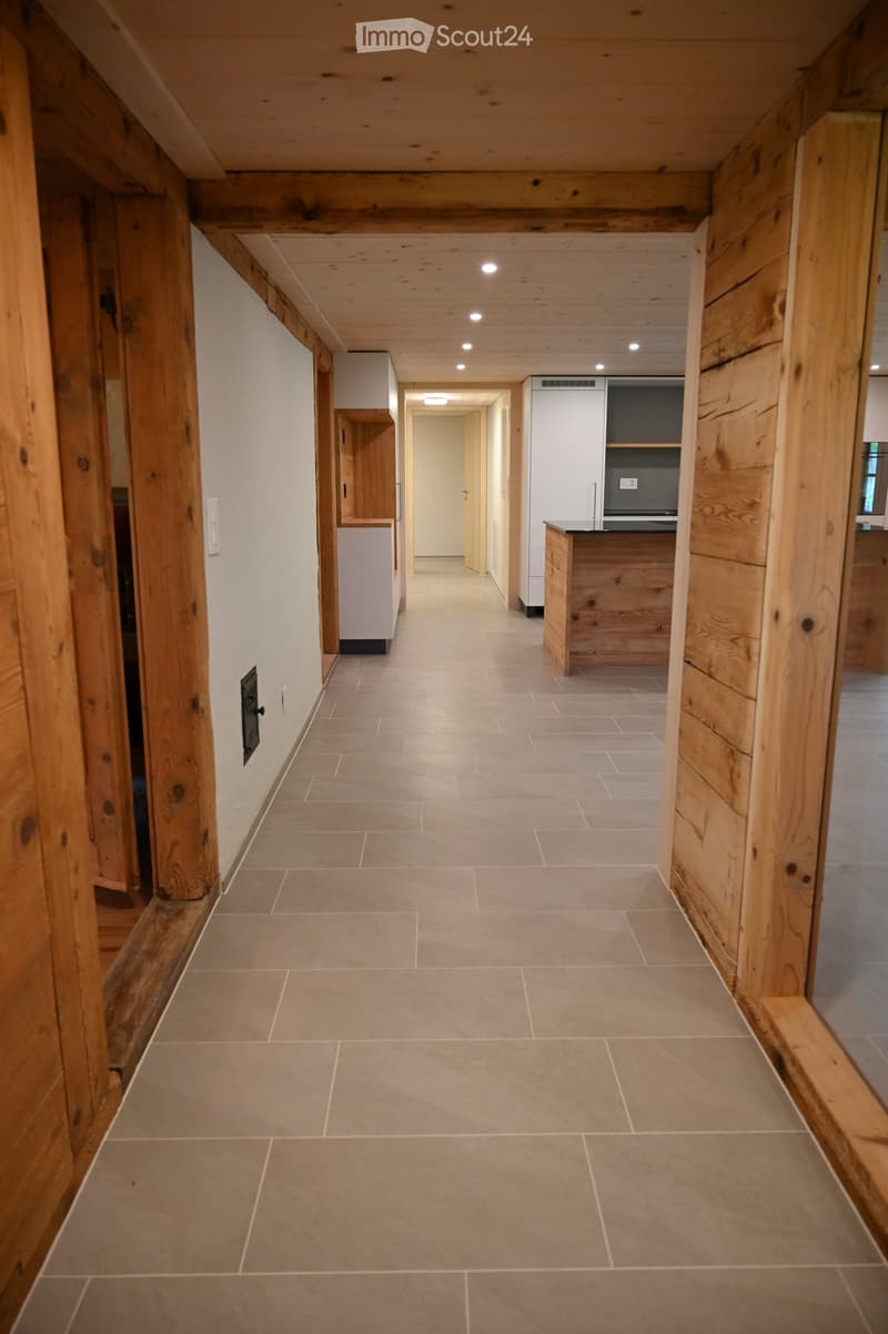 Spacious hallway with wooden beams and tiled floor, leading to a kitchen area with white cabinets and a countertop.