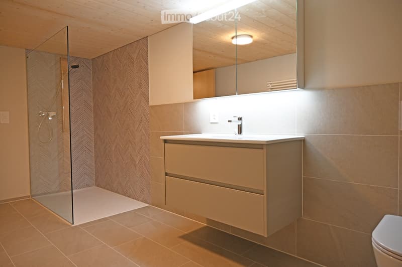 Modern bathroom with a glass-enclosed shower, a sink with a sleek faucet, a large mirror, and a toilet. The space features light wood paneling on the ceiling and walls, complemented by beige tiles on the floor and walls.