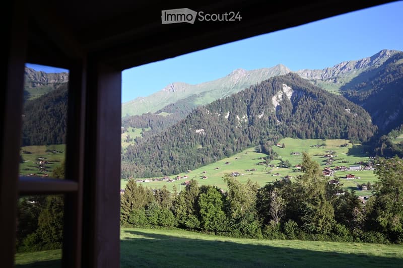 View from a balcony or terrace overlooking a picturesque valley with scattered houses, lush green fields, and distant mountains under a clear blue sky.