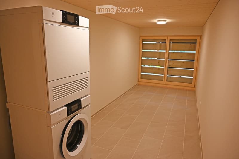 A small, well-lit room with beige tiled flooring and light-colored walls. A modern washing machine and dryer are stacked against one wall, and a large window with blinds allows natural light to enter.
