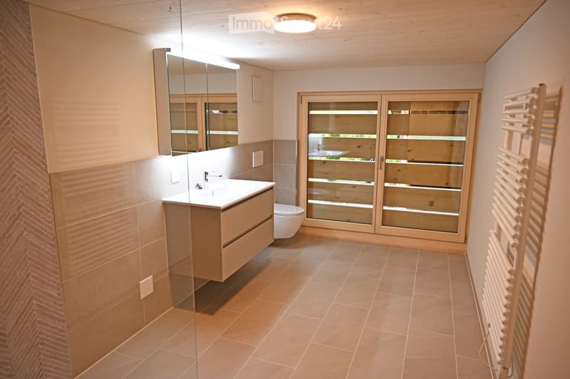 Modern bathroom with a white sink, large mirror, toilet, and a wooden radiator. The room features beige tiles and a large window with wooden frames.