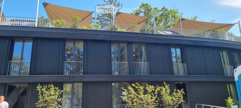 Modern two-story building with dark wooden facade, large windows, and multiple balconies. The roof features a large canopy and additional seating area.