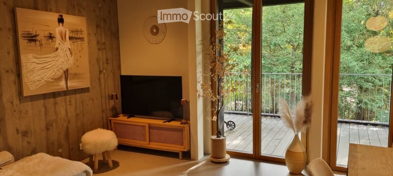 Modern living room with large windows offering a view of greenery, featuring a television on a wooden stand, a small round stool, decorative vases, and a potted plant near the balcony entrance.