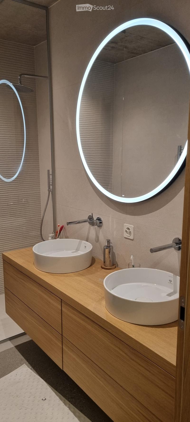 Modern bathroom with two white sinks on a wooden vanity, illuminated round mirror, sleek faucet, and toothbrush holder.