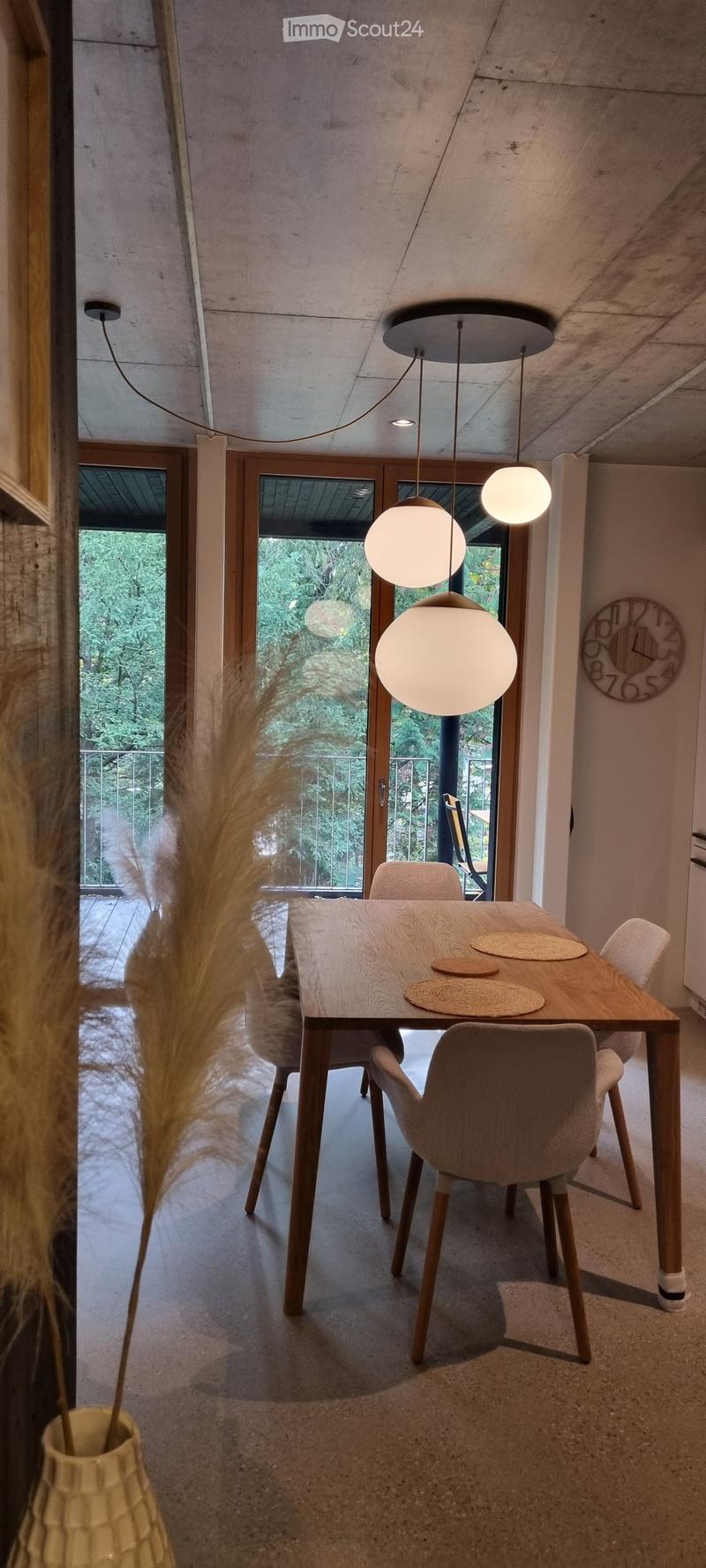 Modern kitchen with a wooden dining table, four chairs, and a decorative vase with tall plants. Large windows and pendant lights are visible.