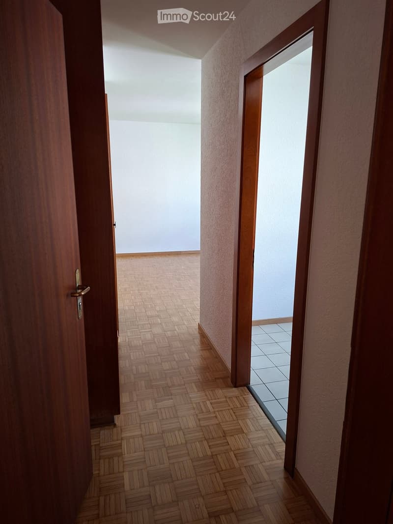 A narrow hallway with wooden herringbone flooring, light-colored walls, and two open doors leading to adjacent rooms.