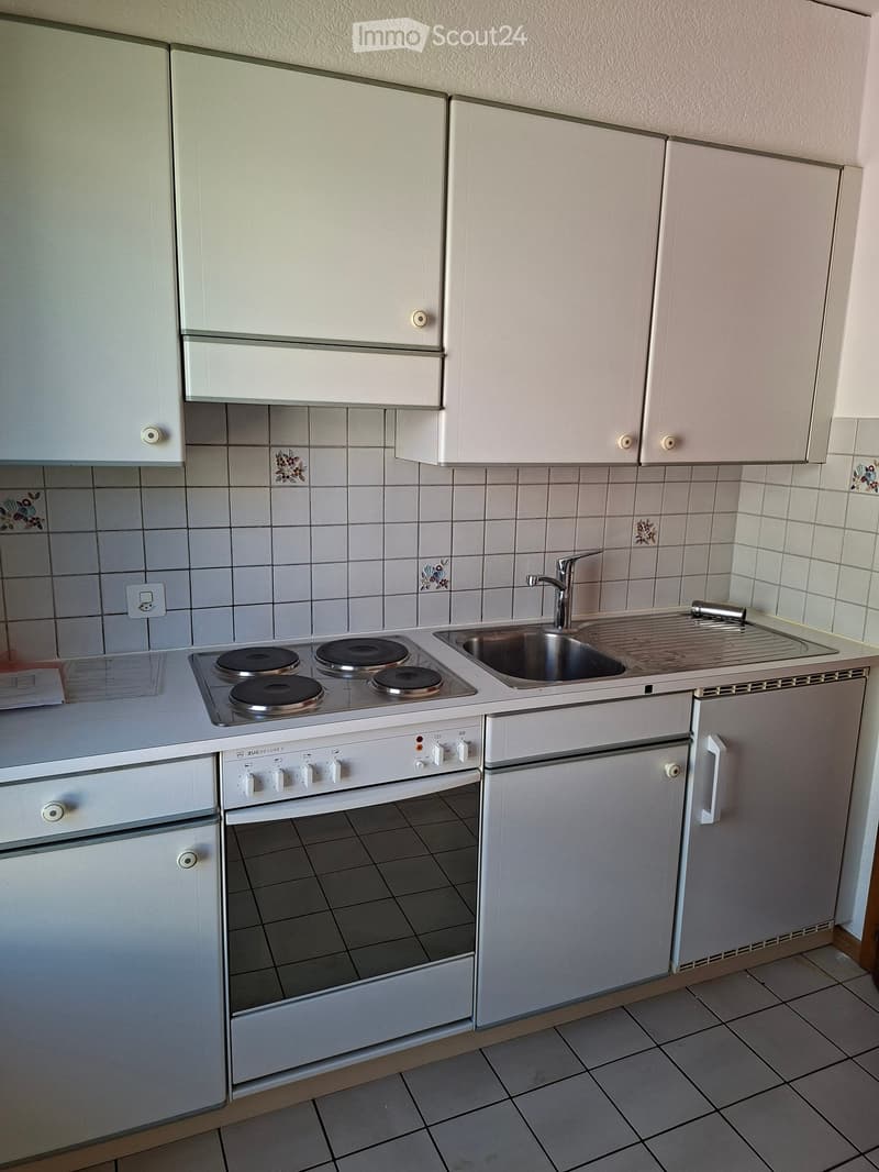 A compact kitchen with white cabinets, a four-burner electric stove, a sink with a faucet, and a refrigerator.