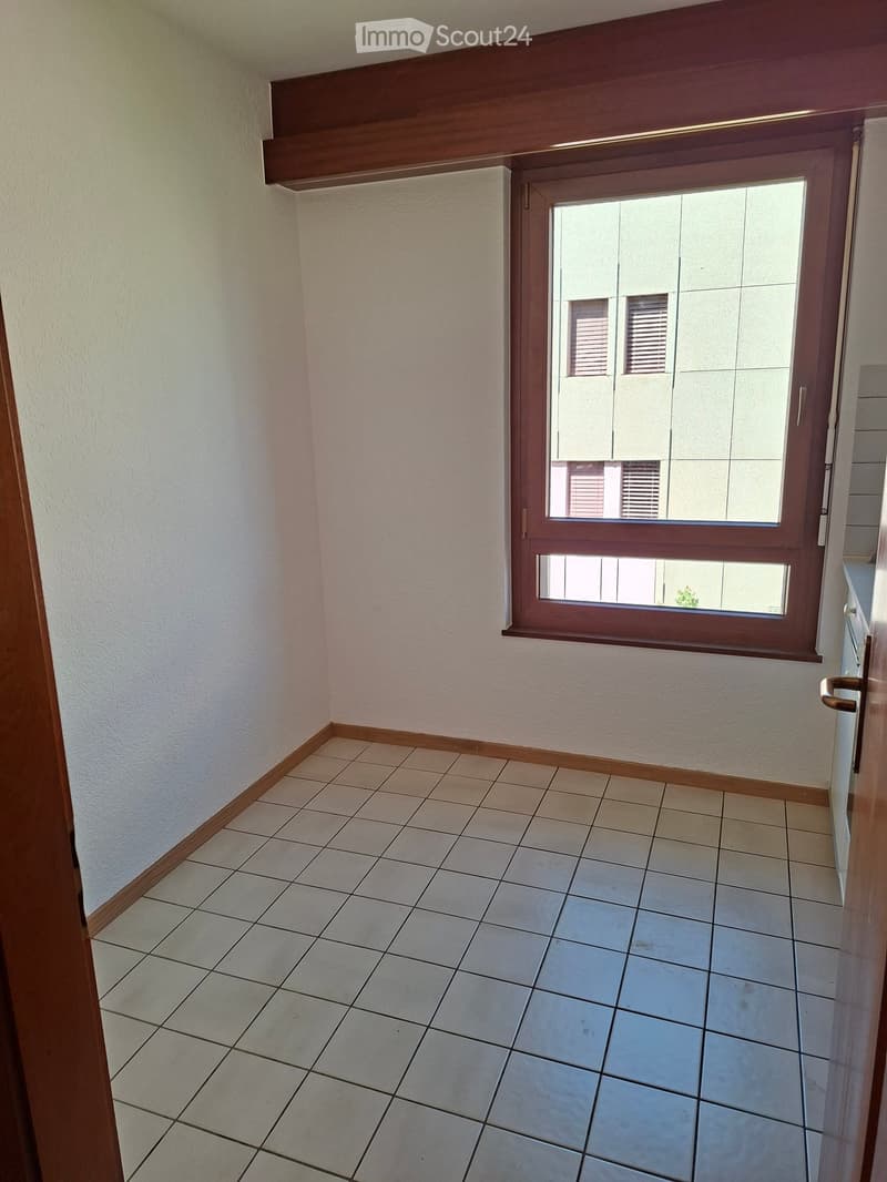 Empty room with tiled floor, white walls, and a large window with wooden frame. The room has wooden baseboards and a visible door frame on the left side.