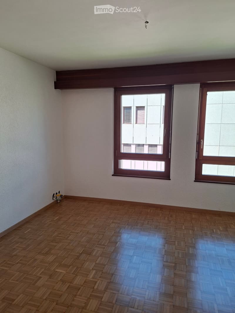 Empty room with wooden herringbone flooring, white walls, and large windows with wooden frames. The room has a simple, clean design with no furniture or decorations.