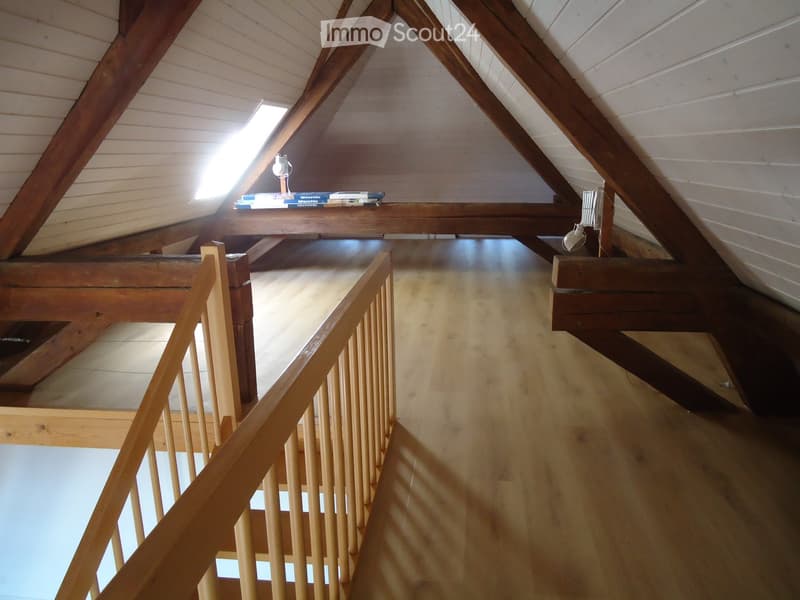 An attic corridor with wooden floors, exposed wooden beams, and a small skylight providing natural light. The corridor features a wooden railing and a small shelf with books and a lamp on the wall.