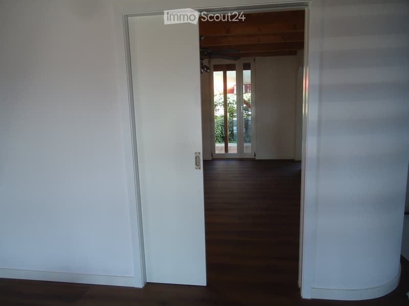 Empty hallway with wooden flooring, white walls, and an open door leading to another room with large glass doors and a view of greenery outside.