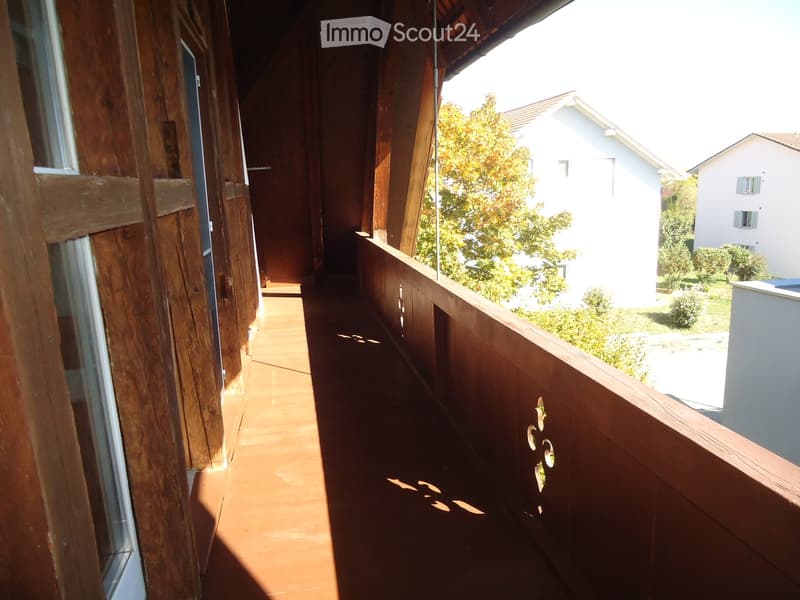 Wooden balcony with terracotta tiles, wooden railing, and view of neighboring houses and greenery.