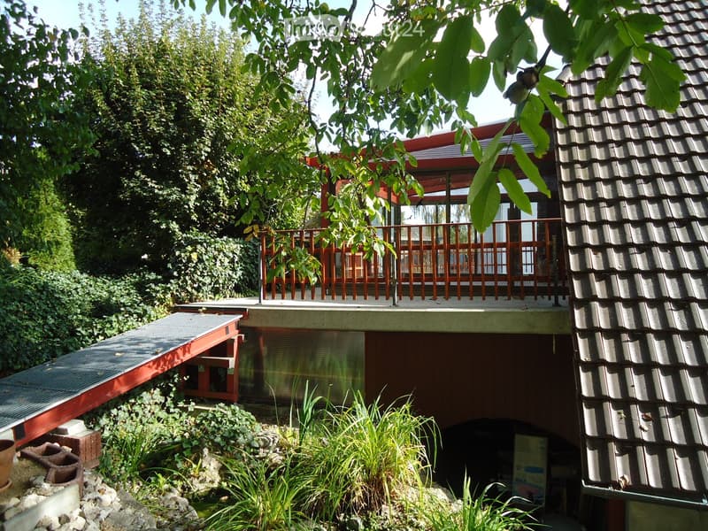 A modern house with a raised ground floor, featuring a wooden balcony, lush greenery, and a small garden area with plants and rocks.