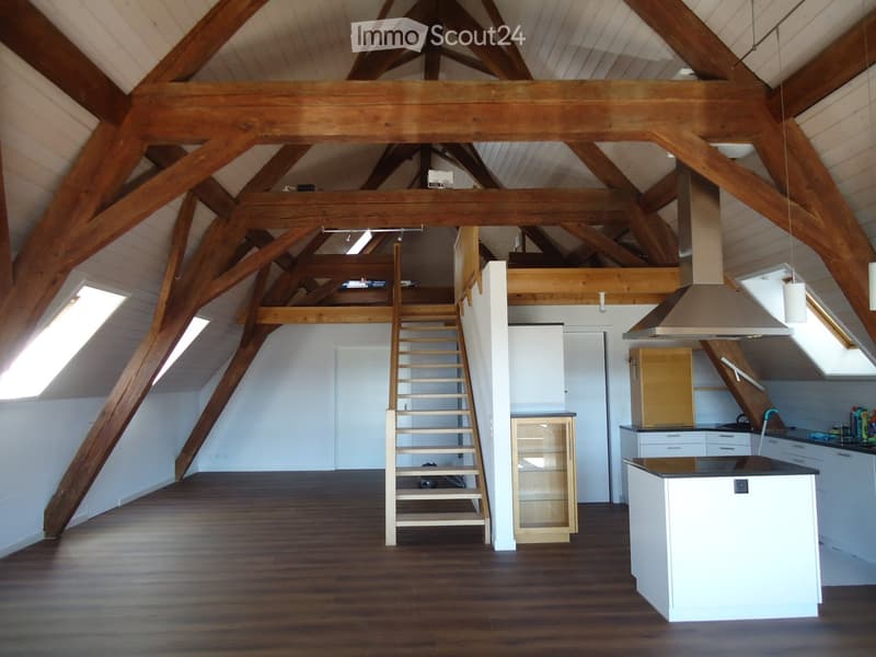 Open-concept attic space with exposed wooden beams, hardwood flooring, a staircase leading to a loft area, a kitchen island, and pendant lights.