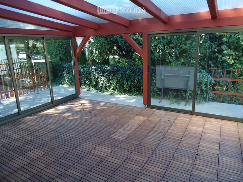 Wooden deck with large sliding glass doors, red-framed roof, and outdoor view of greenery.