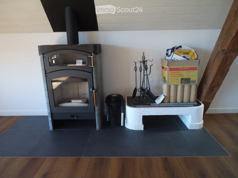 A modern fireplace with a black stove, surrounded by a white base and a set of fireplace tools, with a stack of firewood and a trash bin nearby.