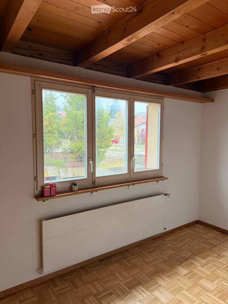 Empty room with wooden herringbone flooring, white walls, large windows with wooden frames, radiator, and a small shelf with a few items on it.