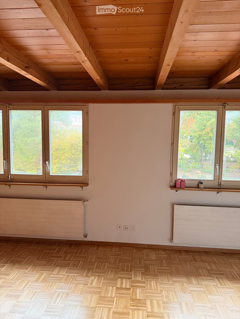 Empty room with wooden herringbone flooring, light-colored walls, large windows with wooden frames, radiators, and exposed wooden ceiling beams.