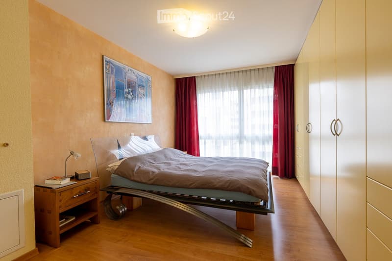 Modern bedroom with wooden flooring, a large bed with a dark duvet, a nightstand with a lamp and books, a tall wardrobe, and a large window with red curtains and a view.