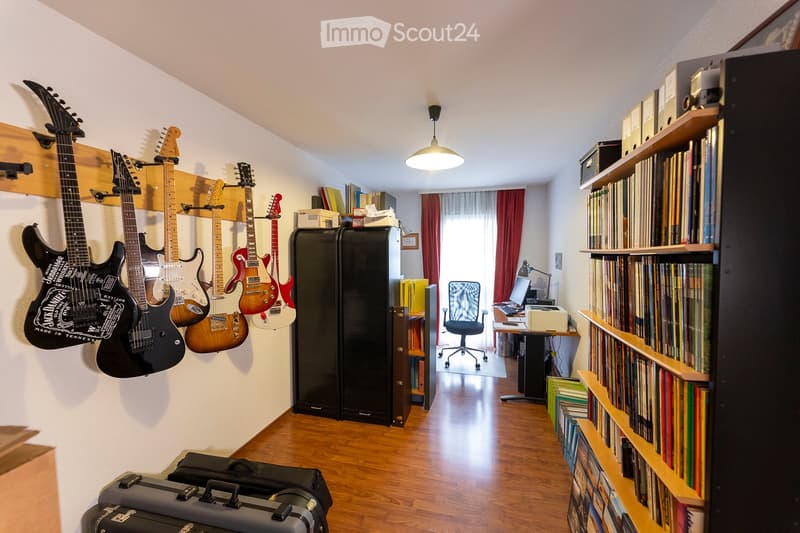 A well-organized room with wooden flooring, multiple guitars mounted on the wall, a bookshelf filled with books and records, a desk with a laptop, a chair, and a hanging lamp.