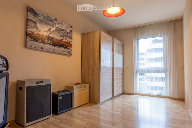 A modern, well-lit room with wooden flooring, a large window with sheer curtains, a wardrobe, and several amplifiers and guitar equipment placed near the wall.
