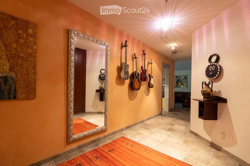 A well-lit hallway with warm orange walls, a large ornate mirror, several guitars mounted on the wall, decorative items, and a patterned rug on the floor.